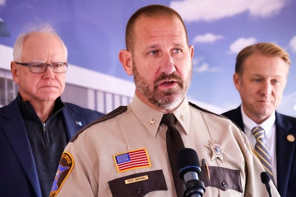 A man in a brown sheriff's uniform speaks into a microphone.