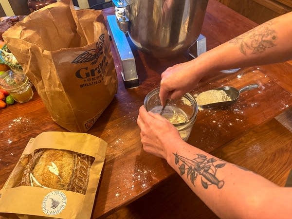 Hands stir ingredients in a glass bowl next to bread and a paper bag on a wooden table.