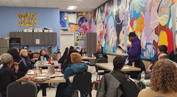 A man in a purple sweatshirt speaks to a room full of people.