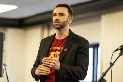 一名男子穿着明尼苏达大学 T 恤和黑色夹克在一次活动中发言。