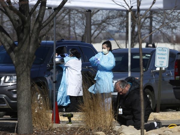 Medical staff wearing safety gear stand outside near cars. 