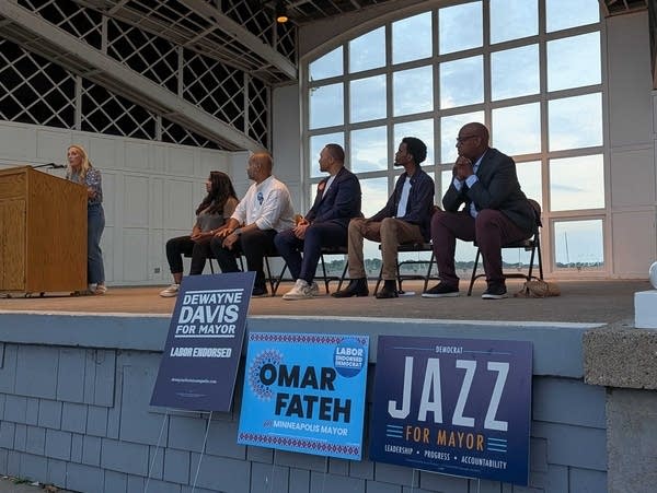 five people on stage listen to a woman at a podium