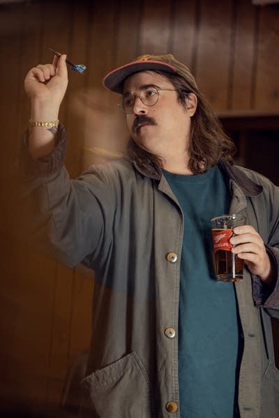 Dan Hornsby wears a baseball cap and barn jacket. He is holding a beer and about to throw a dart.