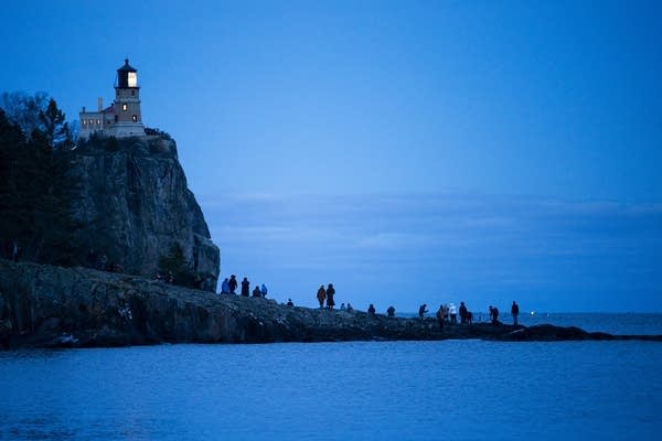 People stand out on a peninsula against a dark blue sky, with a beacon on the cliff nearby.