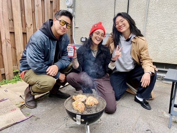 Three people pose for a photo while crouched next to a small grill cooking chicken.