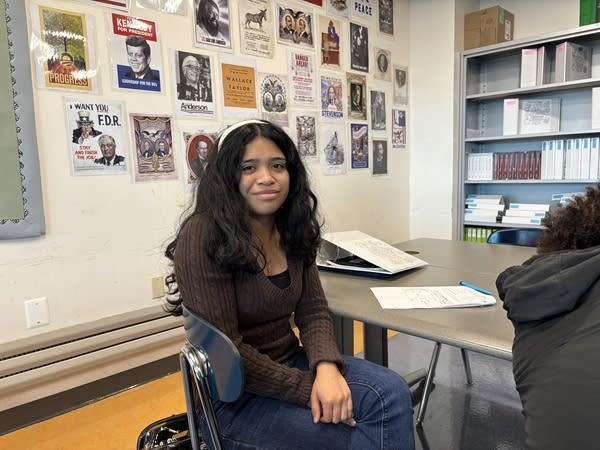 A young woman with black hair and a brown sweater sits in a classroom