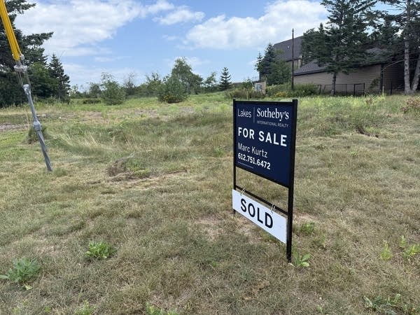 A navy "For Sale" sign stands in a field with "SOLD" hanging from it.
