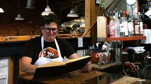 A woman in a white apron leafs through a guest book behind a bar.