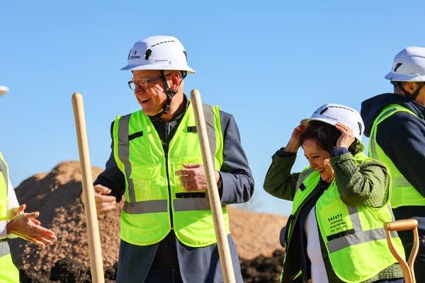 A man and woman in hard hats and green hi-vis vests smile next to shovels.