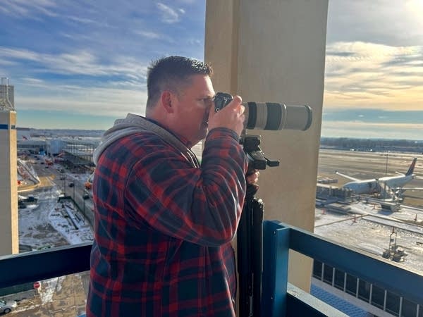 A man looks through a camera's zoom lens toward an airport.