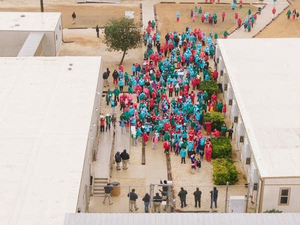 Detainees wearing pink, blue and green jackets gather in the yard of a detention facility.