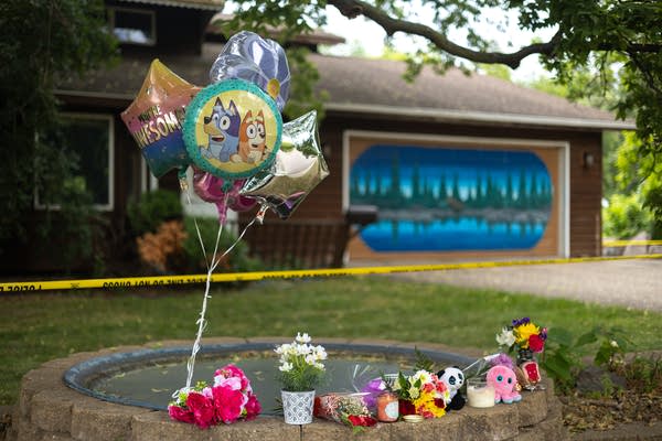 A memorial with balloons, flowers and stuffed animals is seen in the foreground. In the background, a house is blocked by police tape.