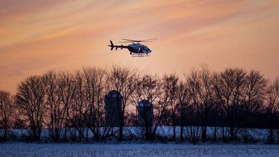 A helicopter hoveres over farm buildings in a snowy field as the sun sets.