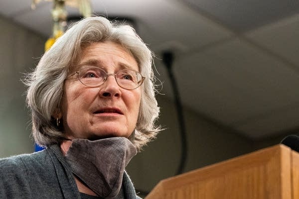 A woman speaks at a podium with mask under her chin.