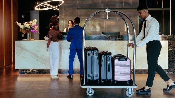 A young hotel Attendant is pushing the luggage cart with suitcases across the lobby while a family is checking into the hotel in the background.