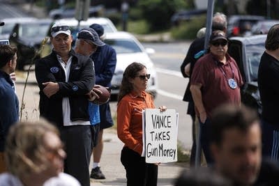 A woman stands in a crowd next to the street. She holds a sign reading "Vance takes gun money."