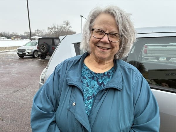 A woman in a blue coat poses for a photo next to a car.