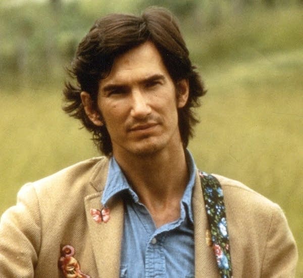 A man with a guitar stands in an open field in the 1970s