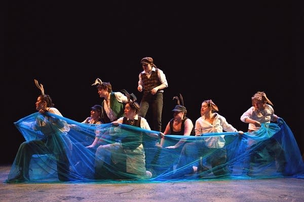 Seven actors stand on a stage, surrounded by a sheen blue cloth depicting water.
