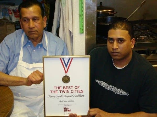 A man in a white apron and a man in a black shirt pose with a framed medal that reads, "The Best of the Twin Cities."