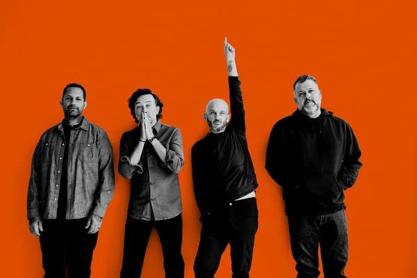 Four men stand in front of an orange background.