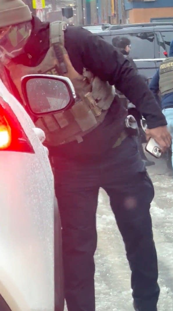 An ICE officer leans into a parked car while holding a cell phone.