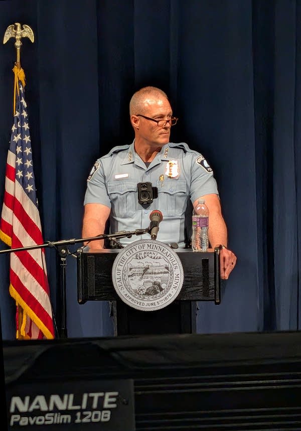 A uniformed police officer speaks at a podium in front of a dark blue curtain while a man in a suit stands next to him.