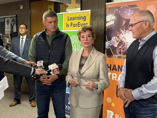 A woman speaks to reporters in a university hallway while flanked by two men.