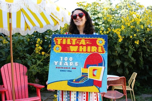 A woman holds a crop art piece that reads "Tilt-A-Whirl."