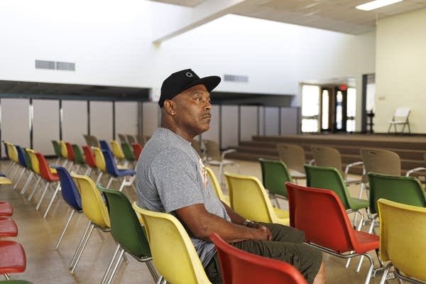A man in a gray T-shirt and black hat sits in a chair.