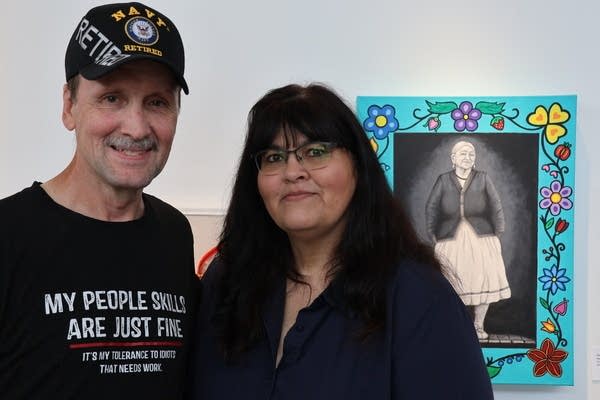 A husband and wife pose for a photo in front of an art piece. It's a black-and-white image of a woman with a bright floral border.