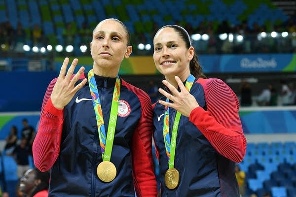 Two women hold up four fingers while wearing gold medals. 