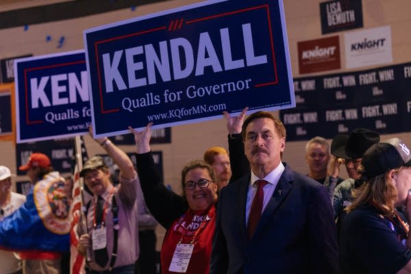 A man in a suit stands under a sign that reads "Kendall Qualls for Governor."