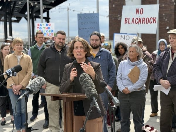 A woman speaks at a podium with a crowd behind her. One person holds a picket sign that reads, "NO BLACKROCK."