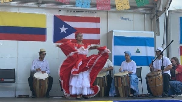 A woman dances with drummers and flags on the stage behind her.