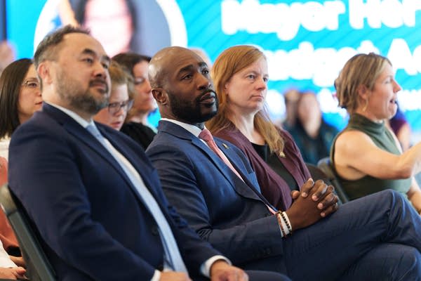 Attendees in suits sit and watch a speech.