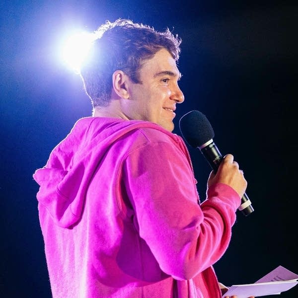 Man in pink hooded sweatshirt standing on a stage and holding a microphone