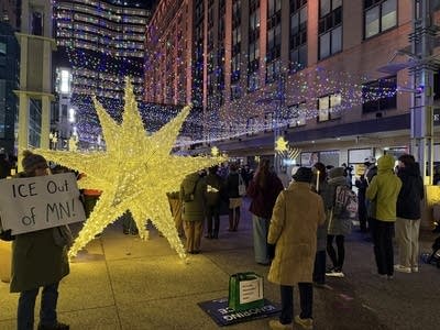 People gather in downtown Rochester's Peace Plaza. A person holds a sign reading, "ICE Out of MN!" in front of a light-up star.