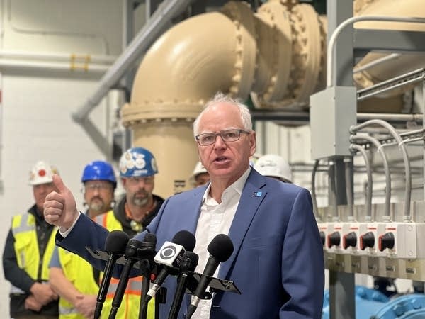 A man stands in front of a stand of microphones, with workers in the background near large water pipes.