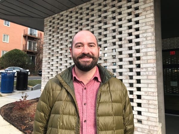 A bald man with a black beard, light red shirt, and green brown jacket smiles and stands outside of a building.
