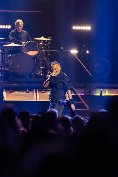 One musician plays drums while another sings onstage during an arena concert