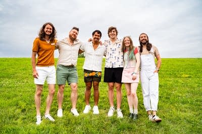 Six musicians stand together on a grassy hillside