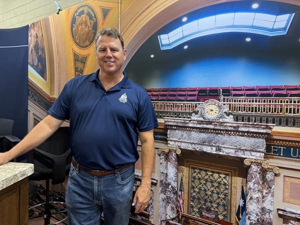 A man in a blue polo shirt poses for a photo in front of a graphic of the state senate chambers.