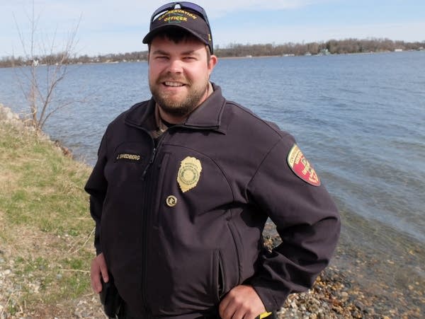 a bearded man wearing a badge stands near a lake