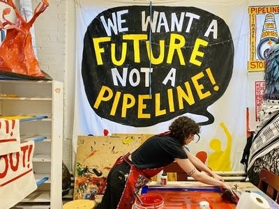 An artist works on a printing, a sign reads "We want a future not a pipeline!" on the wall behind.
