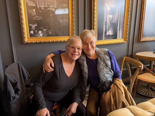 Two women smile for a photo in a restaurant. One has her hand around the other's shoulder.