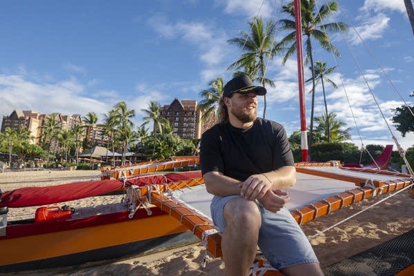A man in a black t-shirt sits on the edge of a boat on a sandy beach.