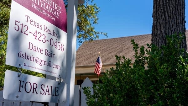 A realtor's "For Sale" sign sits in front of a house covered by trees and bushes.