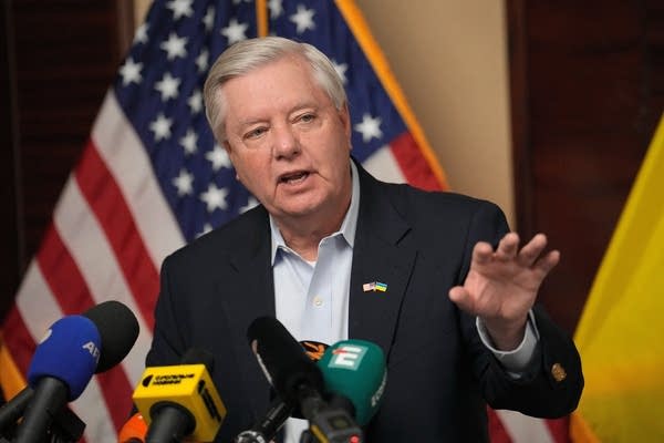A politician in a suit raises his hand while speaking at a podium.