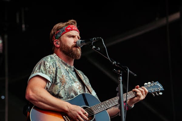 A musician sings and plays guitar on an outdoor stage.